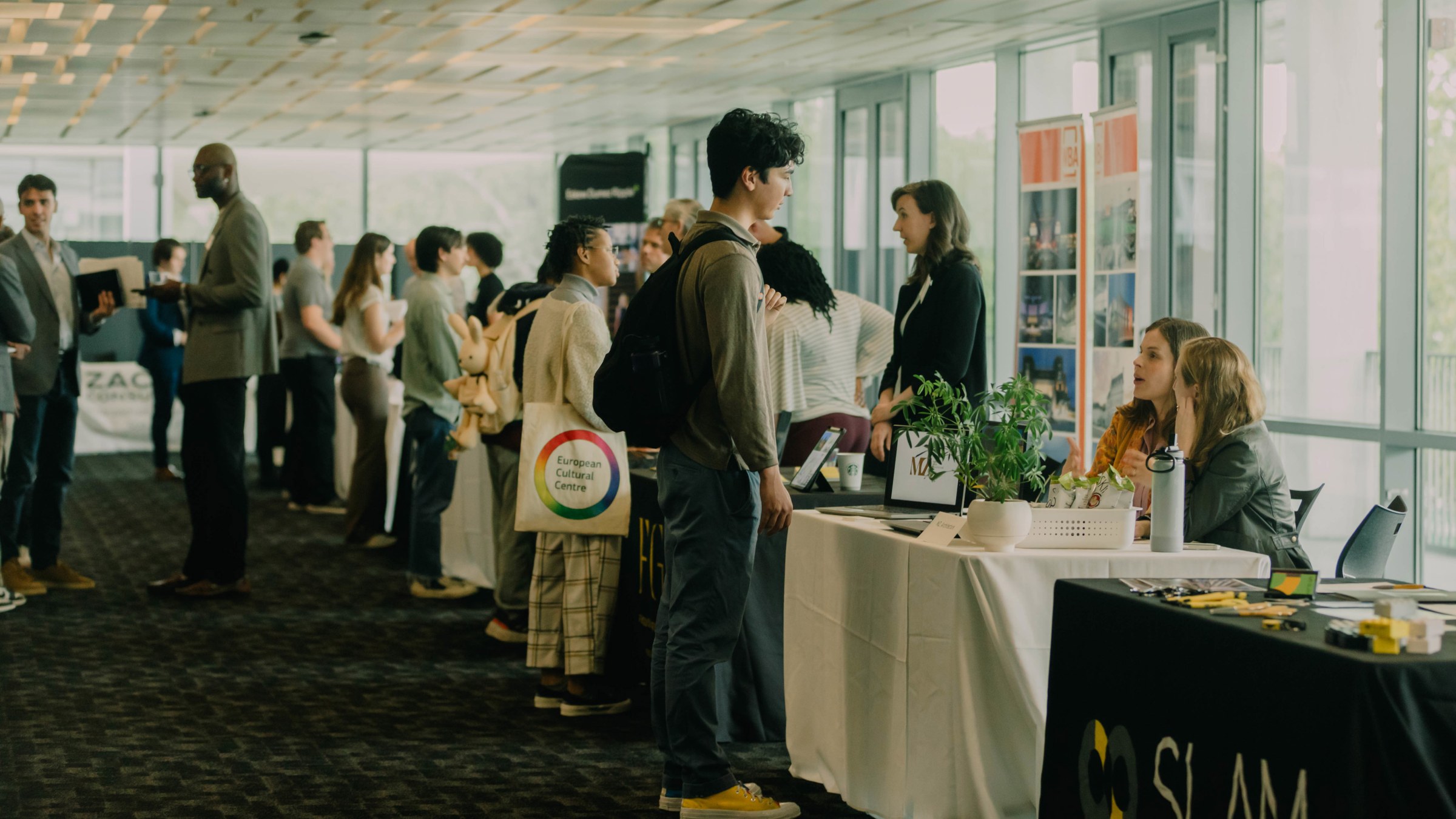 Students stand next to a long row of tables, facing the tables and talking to employers sitting and standing on the other side of the table, inside a large room for a Career Days event.