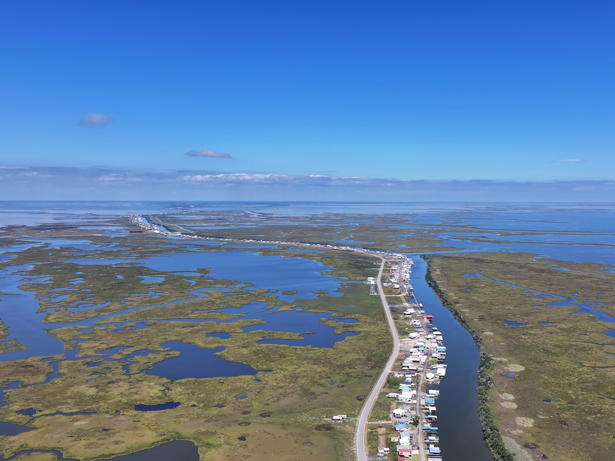 Aerial photo of a winding bayou, the left side lined with a single street of homes surrounded by sparse wetlands stretched out into the horizon.