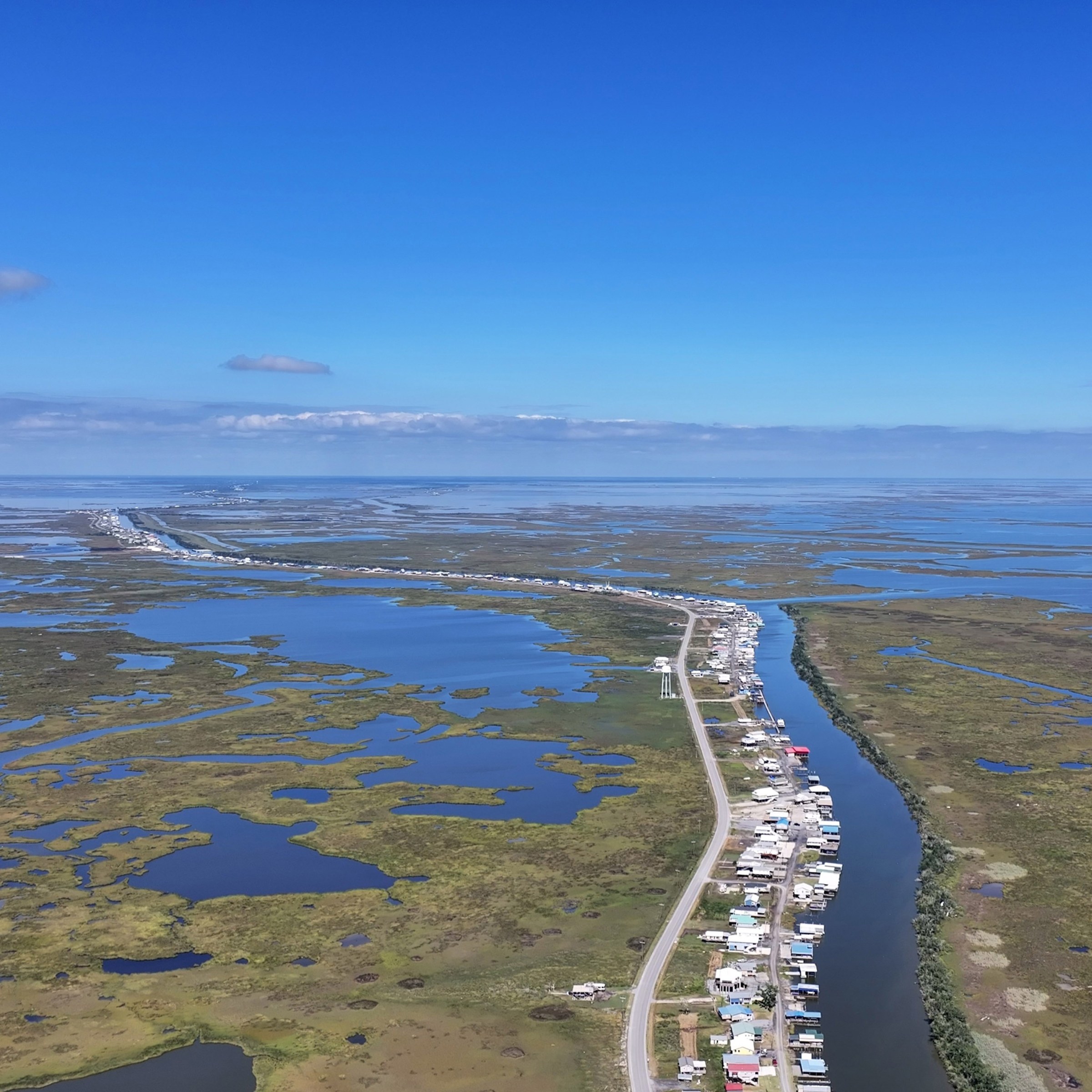 Aerial photo of a winding bayou, the left side lined with a single street of homes surrounded by sparse wetlands stretched out into the horizon.