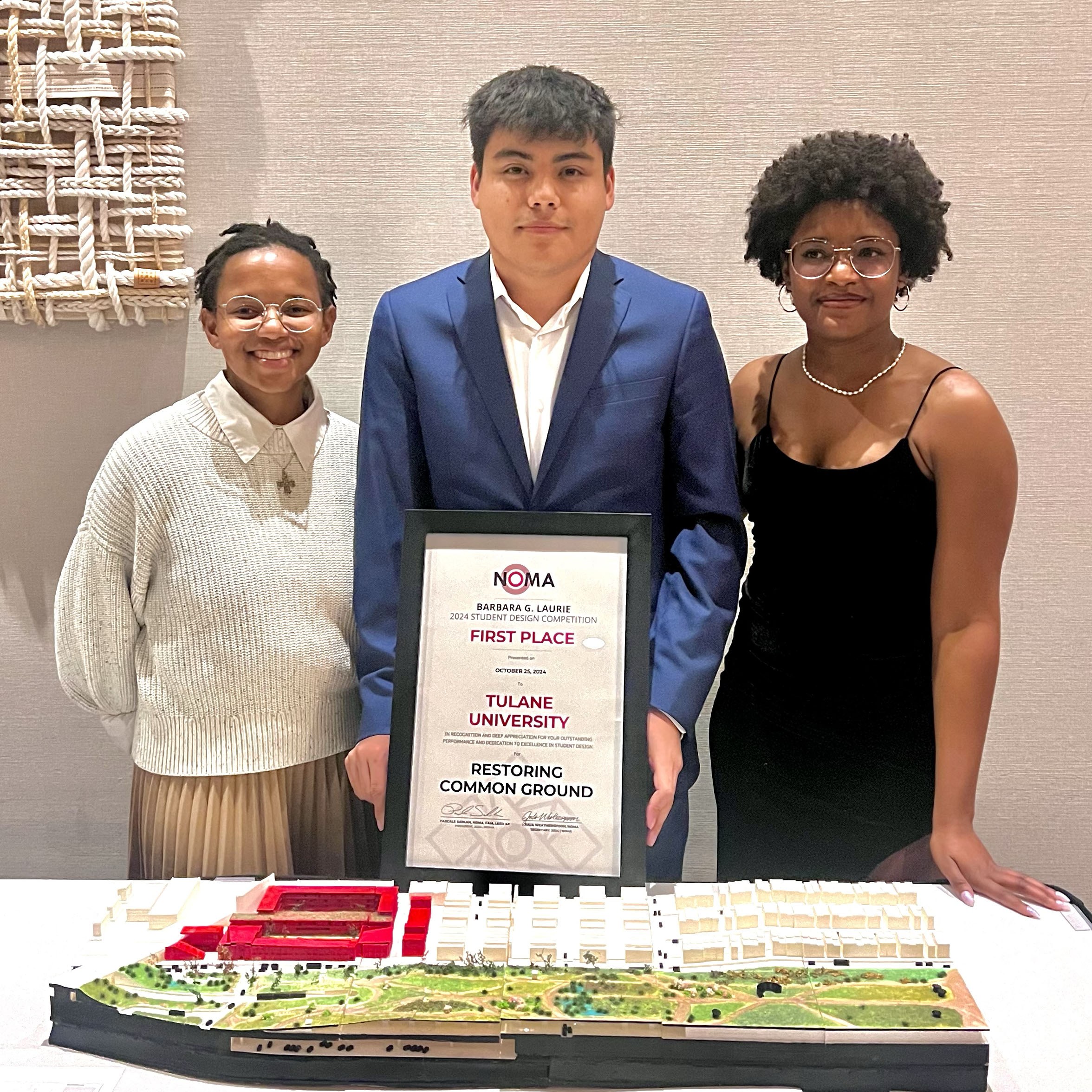 Three students stand behind a table while holding their first place award placard with their model on the table for design "Restoring Common Ground."