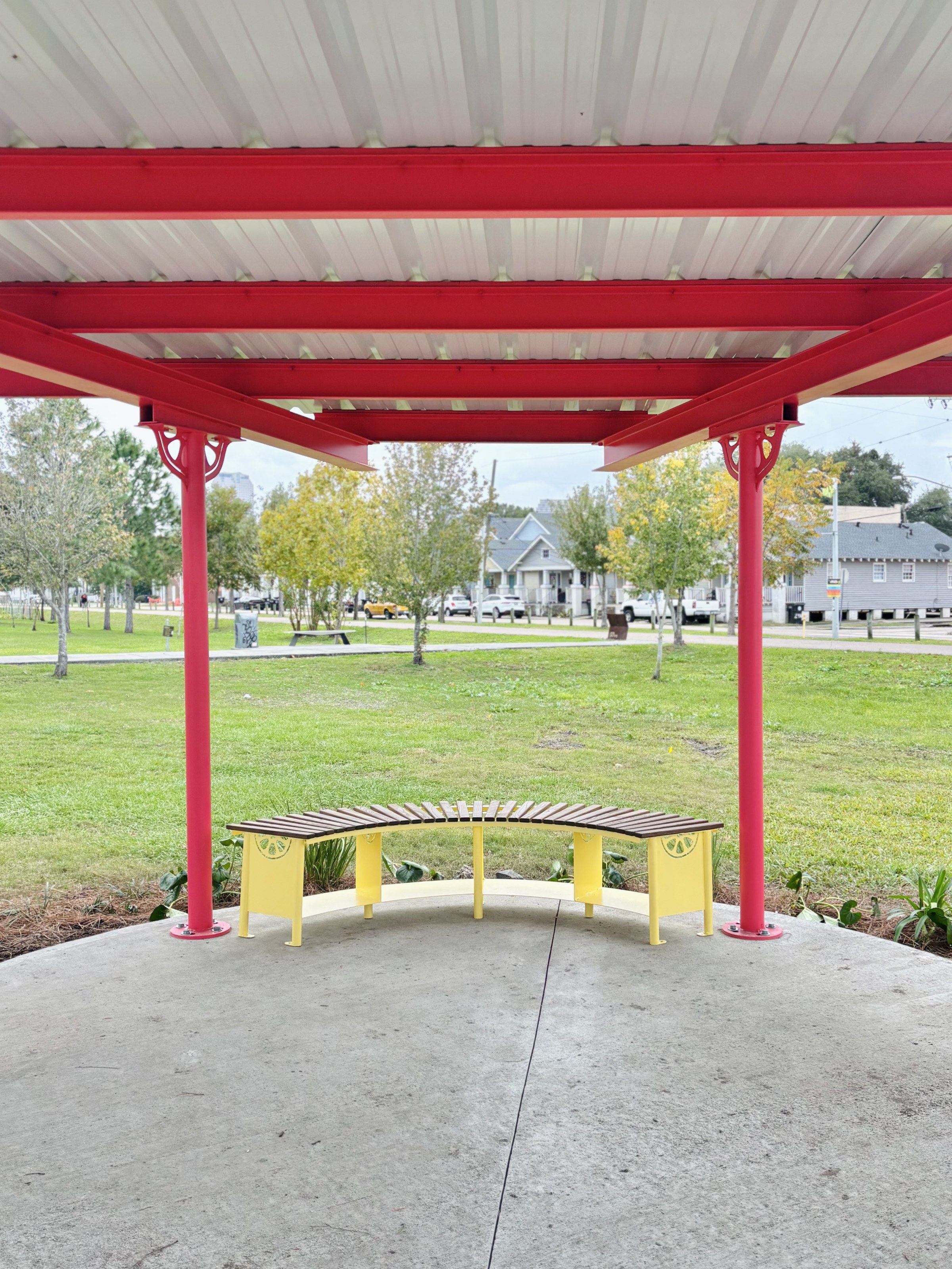 Under the Sprout House shade pavilion, looking from the interior and over a curved bench out to the Lafitte Greenway in the background.