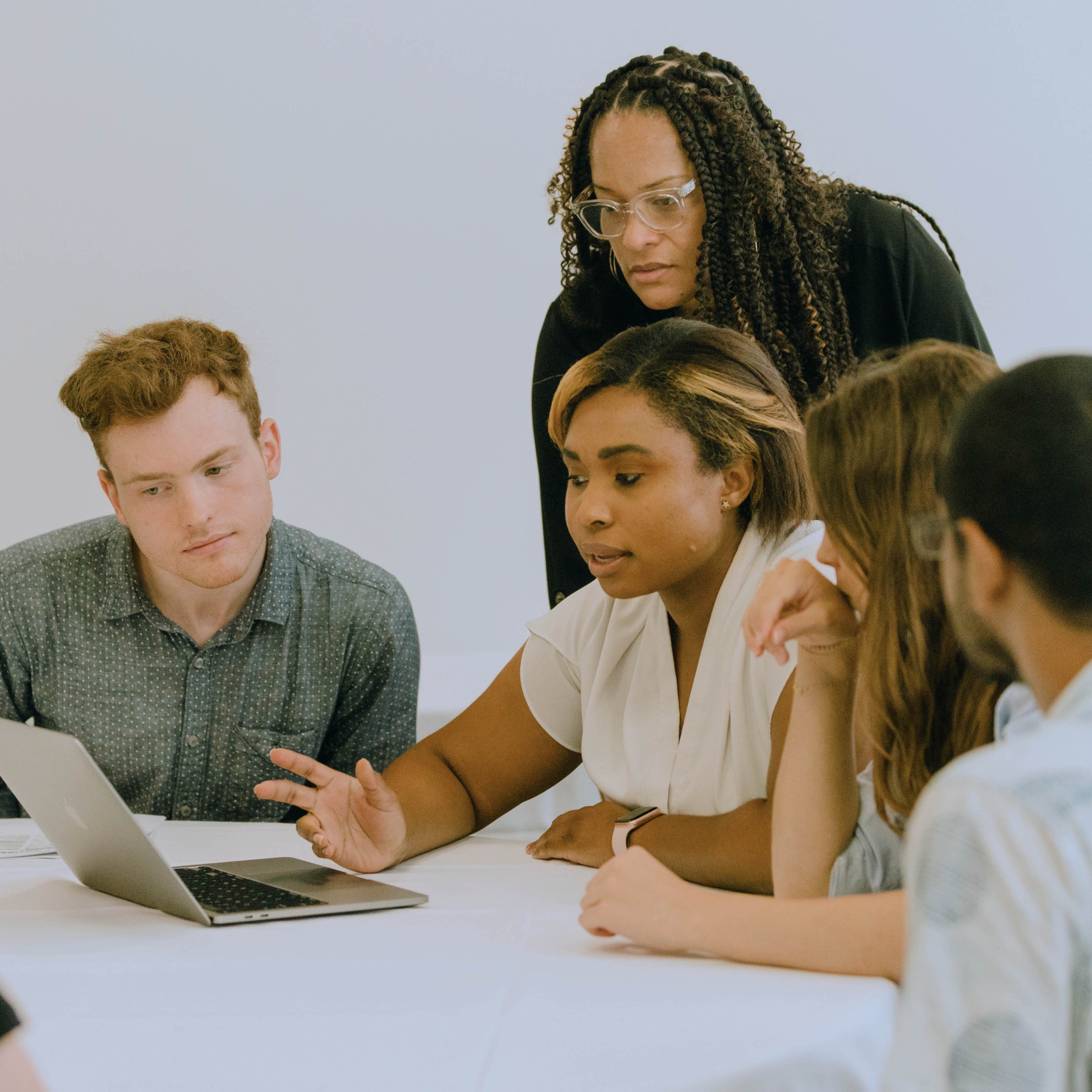 Four students sit at a table looking over a laptop and deep in conversation, while a faculty stands behind them looking intently over their shoulders at the laptop screen.