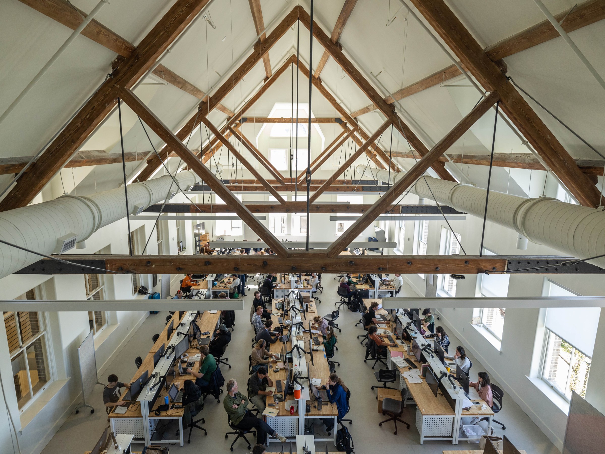 View from above of dozens of students and faculty working at three long rows of desks inside a large studio space with vaulted, pitched ceiling and exposed historic wood beams.