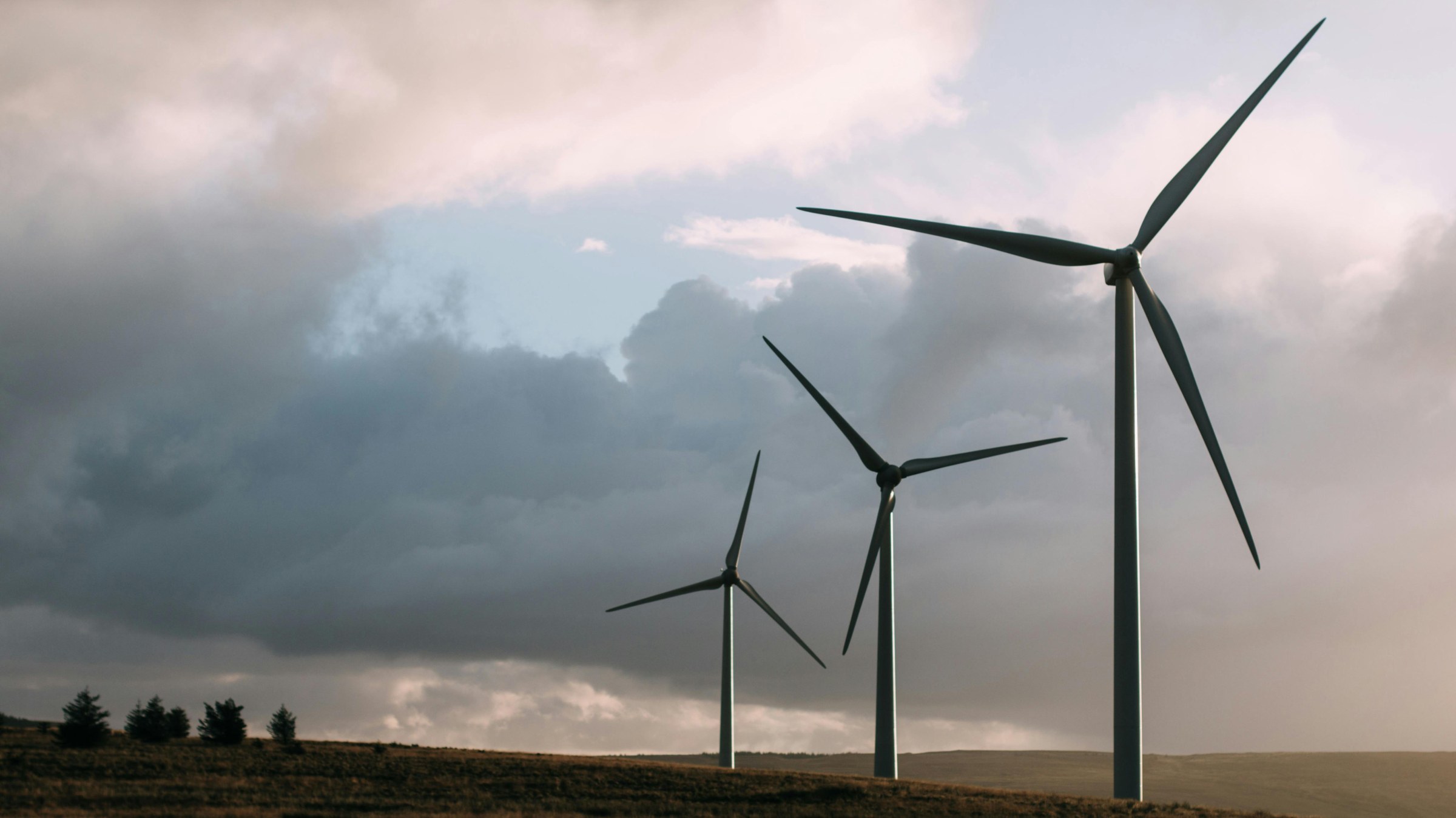 View of three wind turbines in foreground with storm clouds and a rural area in the background.