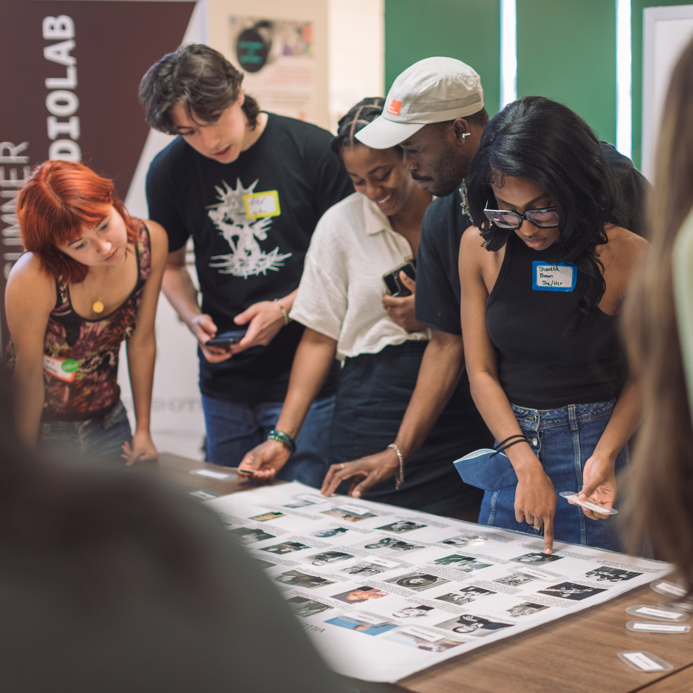 Students stand around a table, while looking down at a poster board with movable images on top.