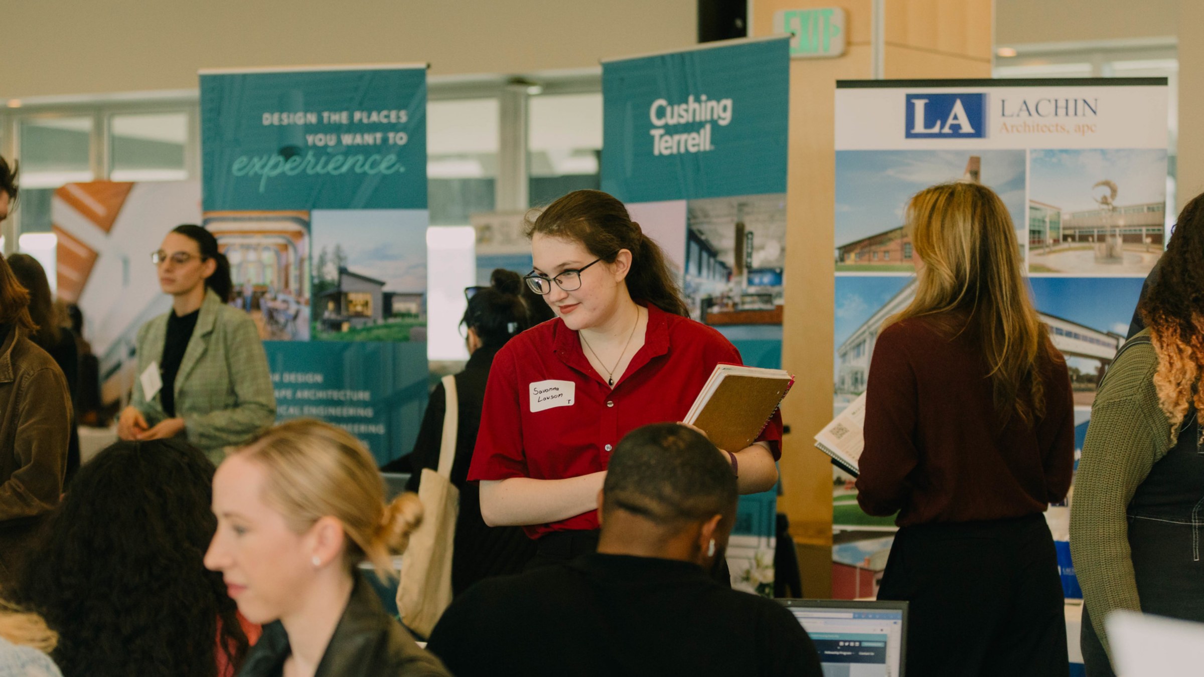 A large room hosting a career fair with rows of employer tables and students walking through the rows.