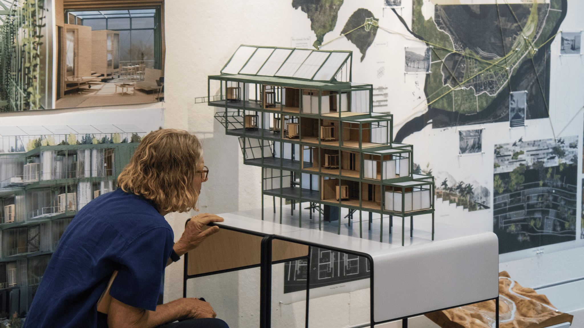 A guest reviewer leans in to closely look at a model of a multi-story, modern building with pinned-up architectural drawings in the background.