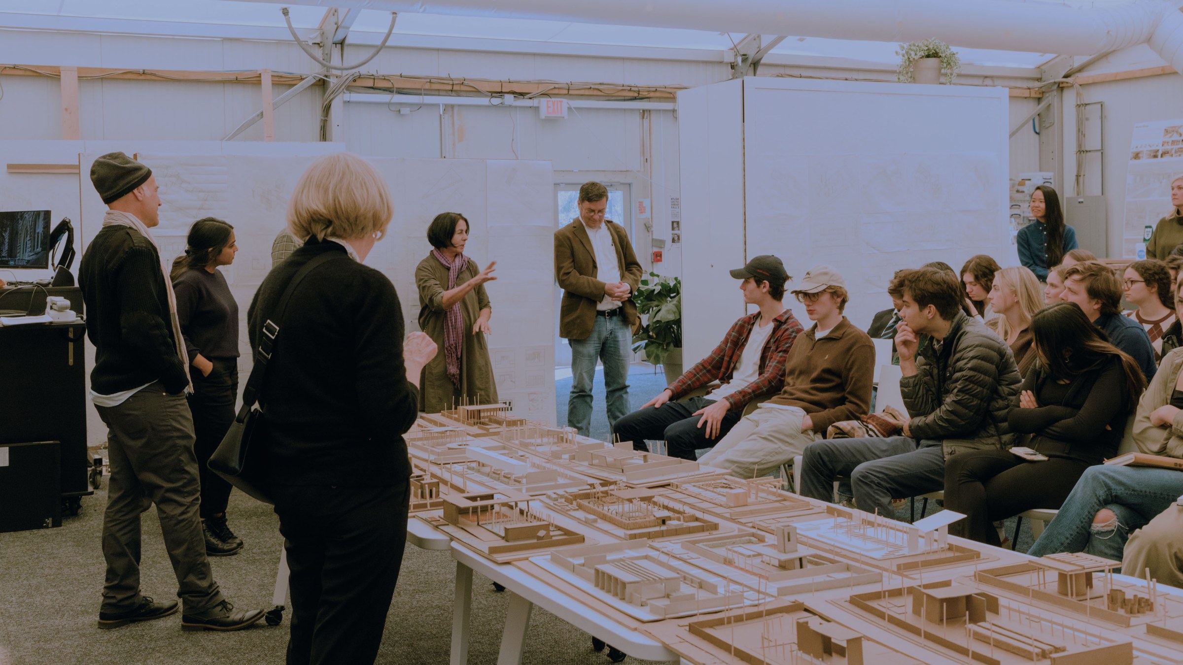 Students sit to the right and faculty stand to the left, looking at a large site model on top of a table in the middle of an open room.