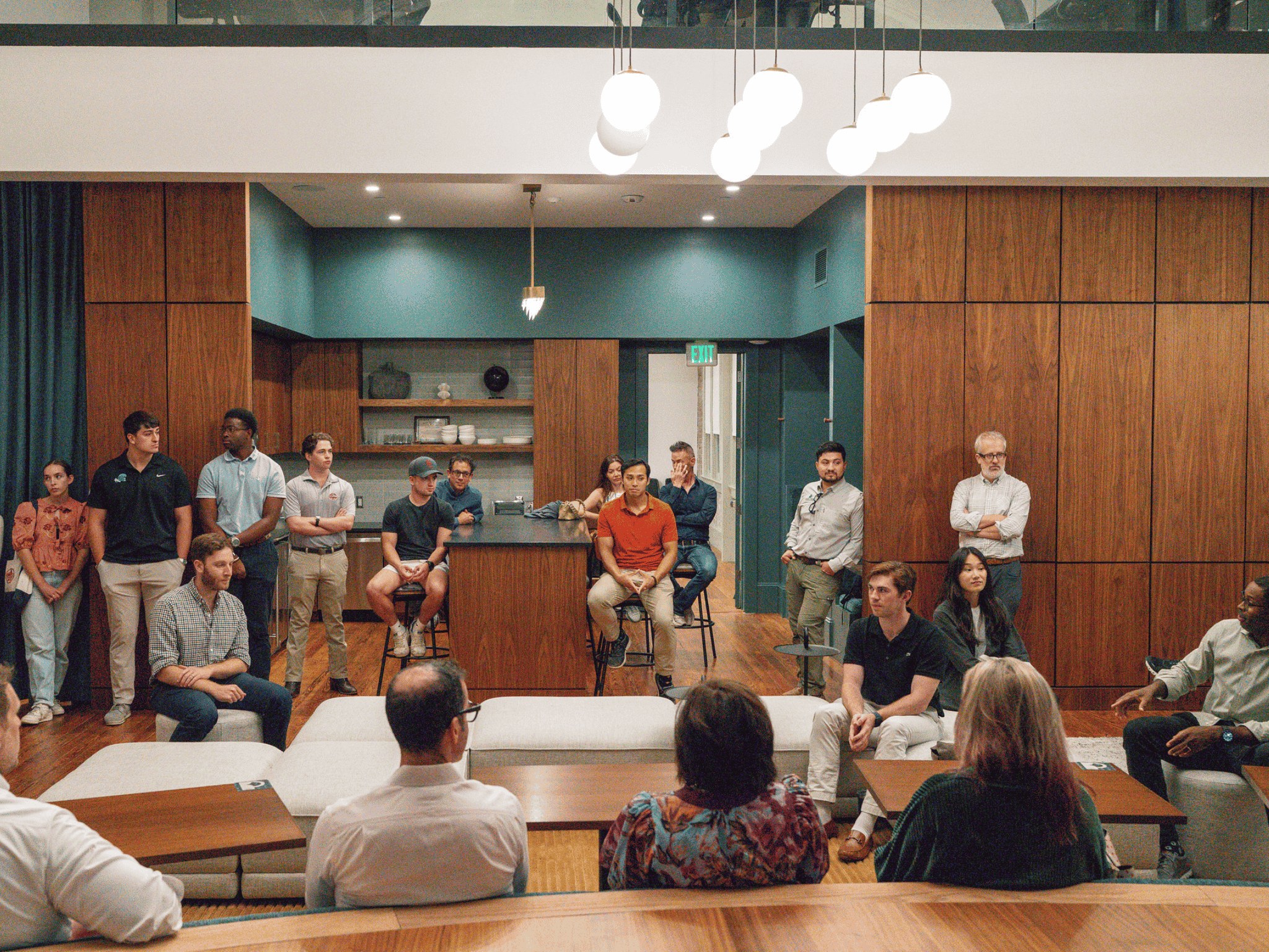 Students and faculty sit and stand while listening to a faculty talk on the right side in an open layout kitchen-dining area of a real estate development project.