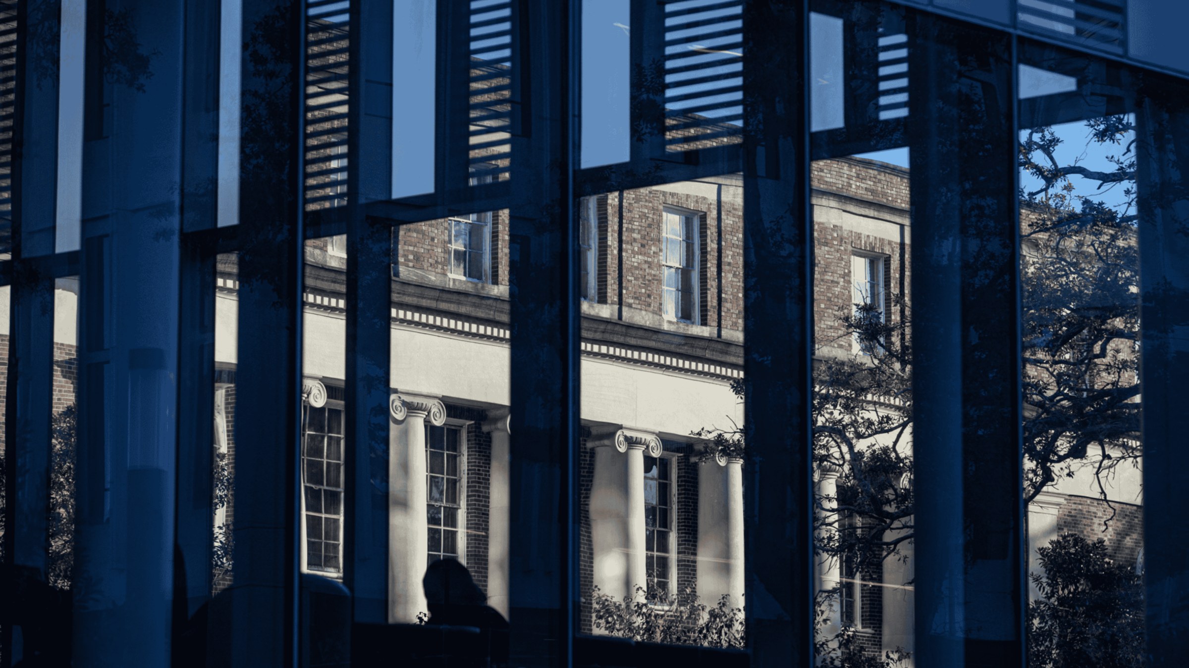 An historic stone building is shown in the angled reflection of a mirror-windowed modern building.